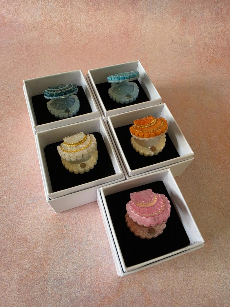 May include: Five small, open shell-shaped trinket boxes in individual white square boxes with black inserts. The shells come in blue, light blue, white with gold trim, orange, and pink, each featuring a textured surface and a small magnetic closure.