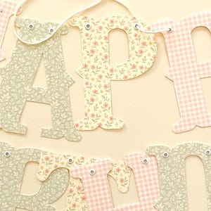May include: A decorative banner spelling out the words Happy Birthday in large, cutout letters. The letters feature alternating patterns of light pink gingham and a sage green floral print, all strung together with a thin white cord through small eyelets.