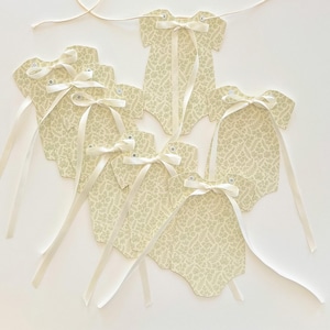 May include: A collection of cream-colored baby bodysuit-shaped tags with a delicate floral pattern. Each tag is adorned with a cream-colored satin ribbon bow and a long ribbon for hanging. The tags are connected by a thin, off-white string.