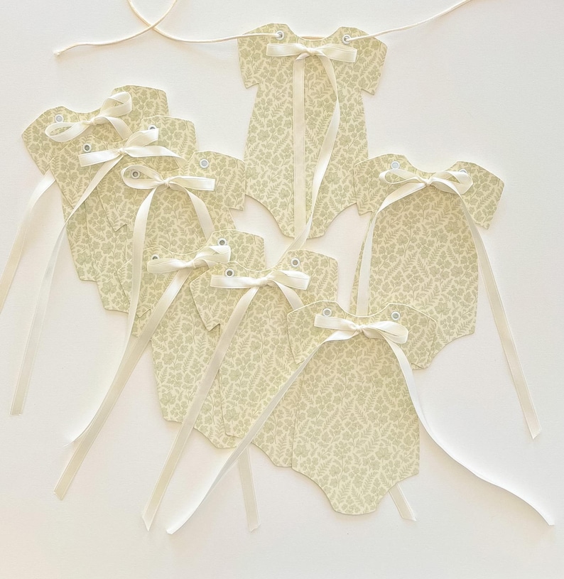 May include: A collection of cream-colored baby bodysuit-shaped tags with a delicate floral pattern. Each tag is adorned with a cream-colored satin ribbon bow and a long ribbon for hanging. The tags are connected by a thin, off-white string.