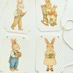 May include: A garland of four decorative tags featuring watercolor illustrations of rabbits. Each tag has a different scene: a rabbit reading, a rabbit reading to another, a rabbit holding gifts, and a rabbit holding a book. The tags are connected by a string.