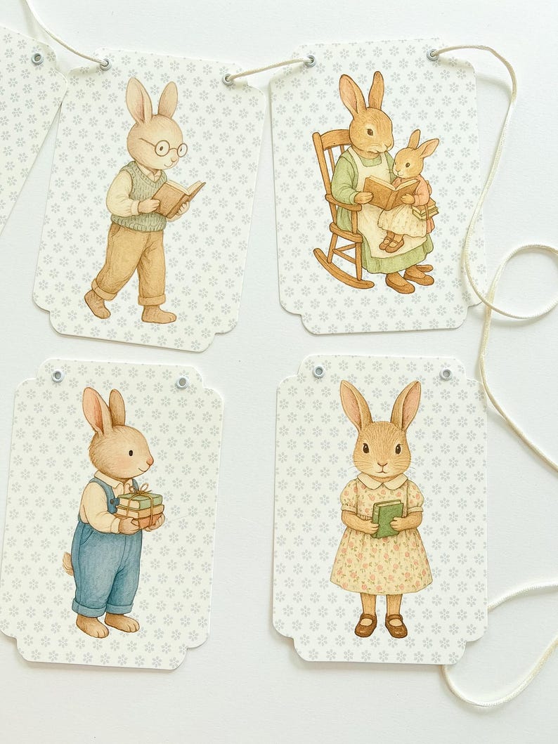 May include: A garland of four decorative tags featuring watercolor illustrations of rabbits. Each tag has a different scene: a rabbit reading, a rabbit reading to another, a rabbit holding gifts, and a rabbit holding a book. The tags are connected by a string.