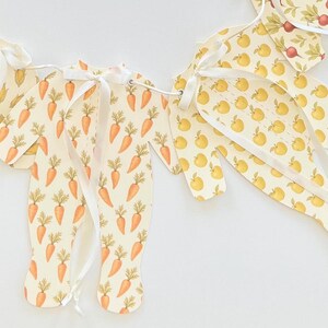 May include: A decorative banner featuring cut-out shapes of baby onesies, apples, corn, and radishes. The onesies are patterned with orange carrots on a cream background. White ribbon is used to connect the shapes.