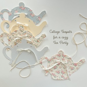 May include: A collection of decorative teapot cutouts in various patterns, including floral and gingham designs. Each teapot is adorned with a white ribbon and strung together with twine. The text "Cottage Teapots for a cozy Tea Party" is also visible.