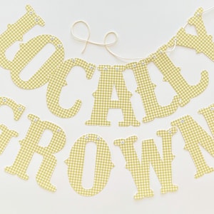 May include: A banner spelling "Grown Locally" in large, yellow and white gingham letters. The letters are strung together with a cream-colored string and have small metal eyelets for hanging. The banner is on a white background.