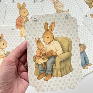 May include: A banner featuring illustrations of rabbits in various scenes. One tag shows a rabbit reading to a smaller rabbit. The background is a light gray with a floral pattern. The banner is strung with a natural-colored cord.