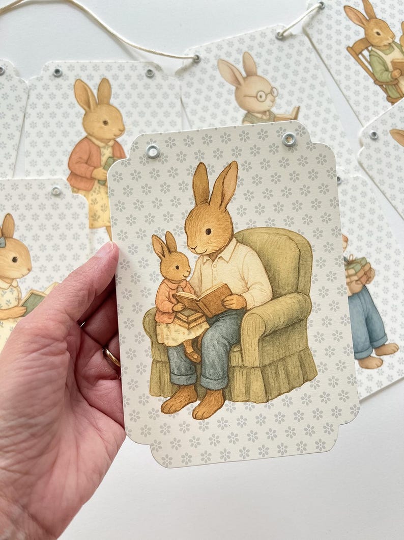 May include: A banner featuring illustrations of rabbits in various scenes. One tag shows a rabbit reading to a smaller rabbit. The background is a light gray with a floral pattern. The banner is strung with a natural-colored cord.