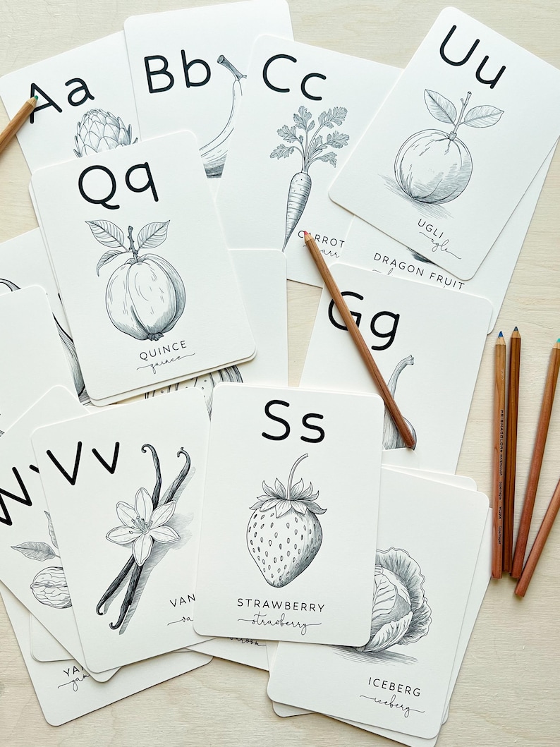 May include: A collection of white flashcards with black illustrations of fruits and vegetables, and the letters of the alphabet. Pencils are scattered around the cards. The cards include the letters A, B, C, Q, U, S, V, and G.