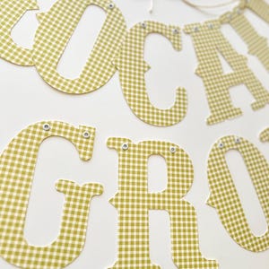 May include: A banner spelling out "LOCAL GROCER" in olive green and white gingham patterned letters. The letters are connected by a thin, natural-colored string. Each letter is individually cut and has small silver eyelets.