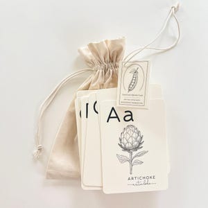 May include: A set of alphabet flashcards with a beige drawstring bag. The cards feature black illustrations of vegetables and the letters of the alphabet. The card for the letter "A" shows an artichoke. The tag reads "Farm Fresh Alphabet Cards".