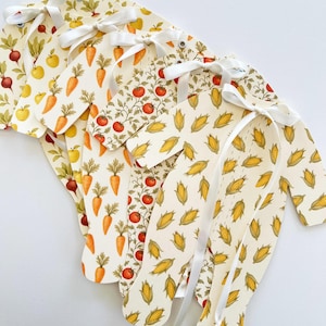 May include: A collection of cream-colored baby onesies with various vegetable patterns, including carrots, tomatoes, and corn. Each onesie is adorned with a white ribbon. The designs are printed on the onesies.