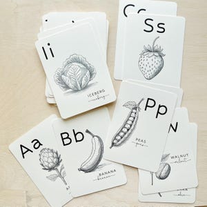 May include: A collection of white flashcards featuring black illustrations of fruits and vegetables. Each card displays a letter of the alphabet and the name of the item. The cards include a cabbage, strawberry, banana, and peas.