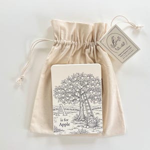 Peut inclure: Un sac en tissu beige contenant des cartes d'alphabet. La carte pr&eacute;sente une illustration en noir et blanc d'un pommier, la lettre "A" et les mots "is for Apple". Une &eacute;tiquette avec une illustration d'oiseau est attach&eacute;e.