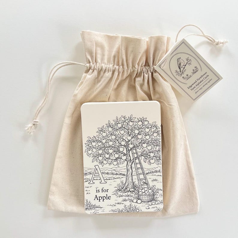 Peut inclure: Un sac en tissu beige contenant des cartes d'alphabet. La carte pr&eacute;sente une illustration en noir et blanc d'un pommier, la lettre "A" et les mots "is for Apple". Une &eacute;tiquette avec une illustration d'oiseau est attach&eacute;e.