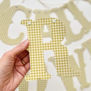 May include: A close-up of a letter "R" from a banner, held in a hand. The letter is a light green and white gingham pattern. Other letters are visible in the background, strung on a white string.