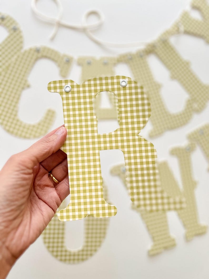 May include: A close-up of a letter "R" from a banner, held in a hand. The letter is a light green and white gingham pattern. Other letters are visible in the background, strung on a white string.