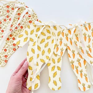 May include: A decorative banner with various food patterns. The banner features sections with tomatoes, corn, and carrots. Each section is connected with white ribbon. The banner is designed with a light cream background.