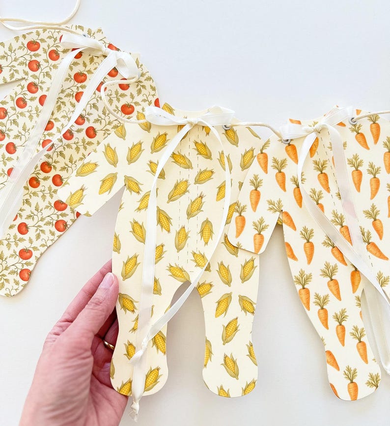 May include: A decorative banner with various food patterns. The banner features sections with tomatoes, corn, and carrots. Each section is connected with white ribbon. The banner is designed with a light cream background.