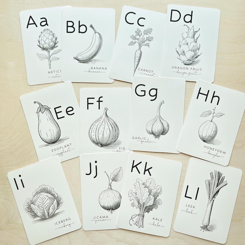May include: A collection of white flashcards featuring black illustrations of fruits and vegetables, each paired with a letter of the alphabet. The cards include an artichoke, banana, carrot, dragon fruit, eggplant, fig, garlic, honeydew, iceberg, jicama, kale, and leek.