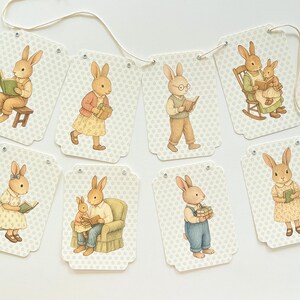 May include: A garland of decorative tags featuring watercolor illustrations of rabbits in various poses. The rabbits are depicted reading books, wearing clothes, and interacting with each other. The tags have a light beige background with a subtle floral pattern.