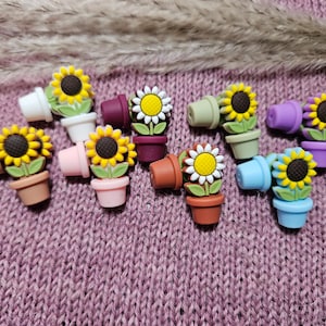 Peut inclure: Collection de breloques miniatures en forme de pots de tournesols de différentes couleurs : jaune, blanc, rose, marron, vert, violet et bleu. Chaque breloque représente un tournesol détaillé avec un centre brun foncé et des feuilles vertes, dans un petit pot couleur terre cuite.