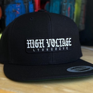 May include: A black and white embroidered patch on a black baseball cap. The patch reads "HIGH VOLTAGE LYNN GROVE".