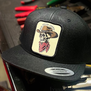 May include: Black snapback hat with a square patch featuring a skull wearing a cowboy hat and a red bandana. The patch reads "BLUE COLLAR COWBOY". The hat is sitting on a toolbox drawer with tools.