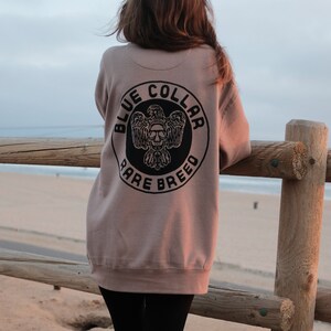 May include: A person wearing a light brown sweatshirt with a black circle logo that says "Blue Collar Rare Breed" on the back. The person is standing in front of a wooden fence with a beach in the background.