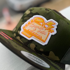 Può includere: Un cappello da camionista verde mimetico con una toppa gialla e bianca che dice "Lineman Hotel" e "We're Here For The Lights On".