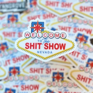 May include: A collection of stickers with a Las Vegas-style sign design. The sign reads "WELCOME to The SHIT SHOW NEVADA" in red, white, and blue. The stickers are in the shape of a diamond with a white border.