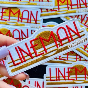 May include: A pile of white patches with the word "LINEMAN" embroidered in red and gold. The patches have a retro design with a sun and stripes.