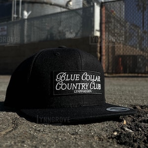 Op de afbeelding: Een zwarte baseballpet met een wit geborduurd patch met de tekst "Blue Collar Country Club - Lynn Grove"