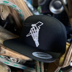 May include: Black snapback hat with a white embroidered design of a skeleton hand holding a lightning bolt. The hat has a flat brim and a structured crown. The hat is surrounded by ropes and leather.