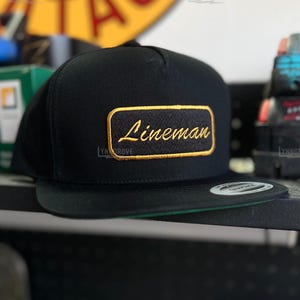 May include: Black flat-brim baseball cap with a green underside. The cap features a rectangular patch with the word "Lineman" embroidered in gold thread. The background includes tools and equipment.