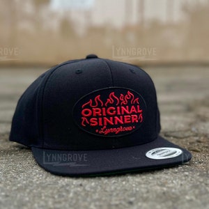 Puede incluir: Gorra de béisbol negra y roja con un parche bordado que dice "Original Sinner" con llamas sobre el texto y "Lyngrove" debajo del texto.