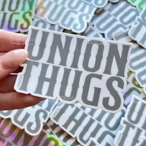 May include: A collection of stickers with the text "UNION THUGS" in a bold, gray font. The stickers have a white border and some have a holographic effect. The stickers are scattered across a surface, with one held in a hand.