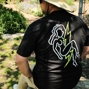 May include: A person wearing a black t-shirt with a white outline of a woman sitting on a lightning bolt. The shirt has the word "LYNNLAKE" printed on the bottom. The person is wearing a straw hat and is sitting on a rock.