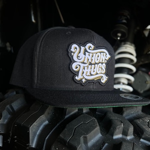 May include: Black and gold baseball cap with a white embroidered logo that says "Union Thugs" on the front.