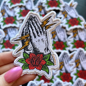 Peut inclure: Patch brodé blanc avec une paire de mains en prière, des éclairs et une rose rouge. Le patch est entouré d'autres patchs similaires.