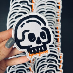 Peut inclure: Une pile de patchs de crâne blancs avec des contours noirs et des coutures orange. Les patchs ont le texte "Lynyrd Skynyrd" brodé en orange sur le bas.