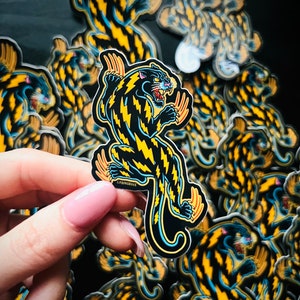 May include: A black panther sticker with yellow and blue lightning bolt patterns. The sticker is in a hand, and there are many other panther stickers in the background.