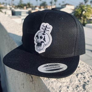 Può includere: Cappellino da baseball nero a tinta unita con visiera bianca e un motivo di teschio bianco e nero. Il cappello ha un'etichetta bianca con la scritta "The Authentic Snapback Yupoong Since 1974".