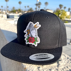 May include: Black and white praying hands with lightning bolts and a red rose patch on a black fitted baseball cap. The cap has a white label that reads "The Authentic Yupoong Snapback Yupoong Since 1974".