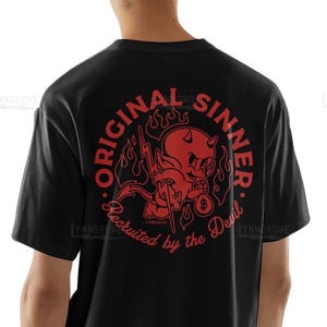 Puede incluir: Camiseta negra con un gráfico circular rojo que presenta un diablo de dibujos animados sosteniendo un rayo y una bolsa de dinero. El texto alrededor del gráfico dice "Original Sinner. Recruited by the Devil."