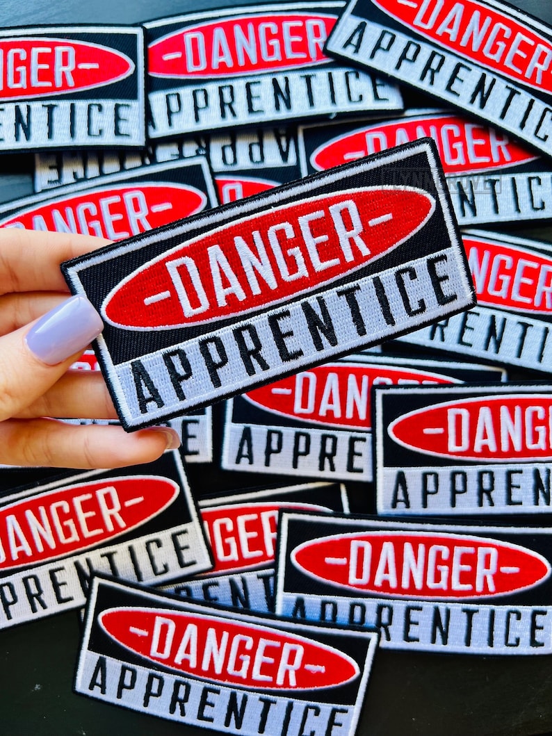 Puede incluir: Primer plano de varios parches negros y blancos con se&ntilde;ales de advertencia ovaladas rojas que dicen "Peligro-Aprendiz".