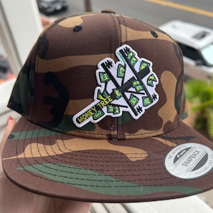Op de afbeelding: Een camouflage baseballpet met een zwart en groen geborduurd patch waarop "Money Tree" staat en een ontwerp van dollarbiljetten. De pet heeft een zwarte klep en een wit "Snapback" label.
