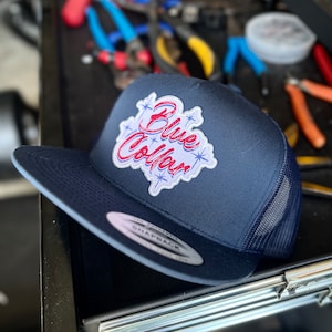 May include: Navy blue trucker hat with a mesh back and a flat brim. The hat features a patch with the words "Blue Collar" in red and white, surrounded by blue starbursts. The hat has a snapback closure.