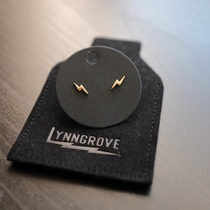 May include: A pair of gold lightning bolt stud earrings displayed on a black velvet jewelry card with the text "LYNNGROVE" printed on the bottom.