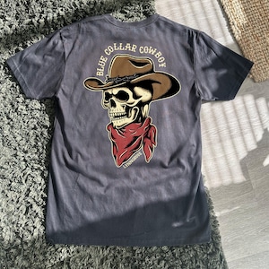 Op de afbeelding: Een donkergrijs T-shirt met een schedel die een cowboyhoed en een bandana draagt. De tekst "Blue Collar Cowboy" staat op de achterkant van het shirt.