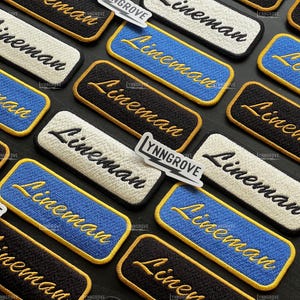 May include: Embroidered patches with the word "Lineman" in a cursive font. The patches are rectangular with rounded corners and come in various colors, including black, white, and blue, with gold or yellow stitching. A small sticker with "YNNGROVE" is also present.